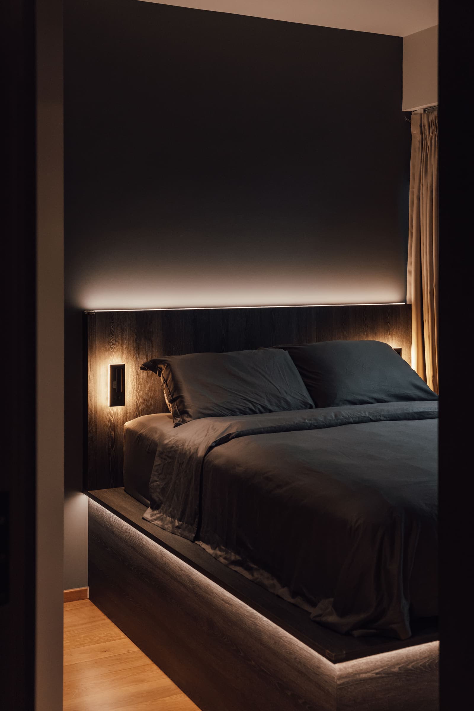Bedroom lit low, with a concealed strip washing the headboard wall and a slim reading light beside the bed.
