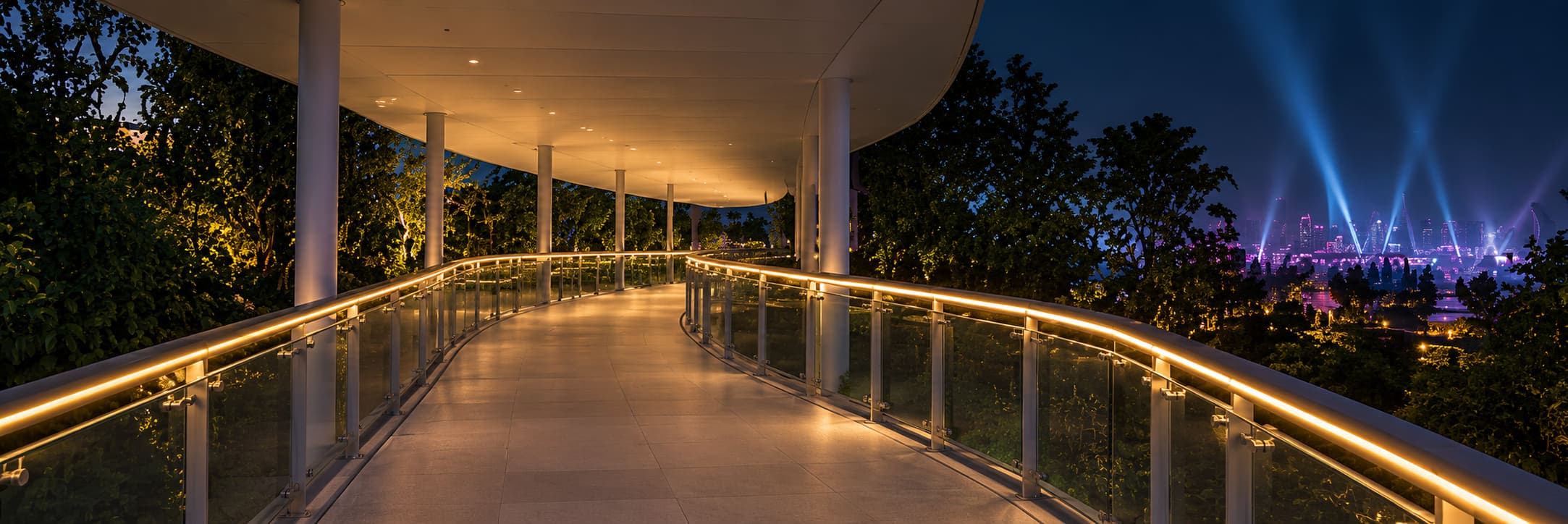 Elevated walkway through planting at night, a continuous warm line along both handrails leading the path, a lit show in the trees beyond.